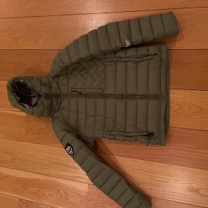 Grön dunjacka från Black Crows - Säljer en snygg grön dunjacka från Black Crows. Jackan har en quiltad design med dragkedja och en cool mönstrad insida. Perfekt för kyliga dagar med sin långa ärmar och stilrena look.