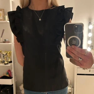 Svart blus med volangdetaljer - Snygg svart blus med volangdetaljer längs ärmarna💘Blusen är ärmlös och har en knapp i ryggen! Okänt märke