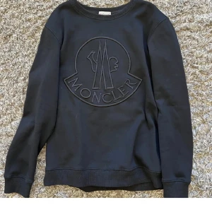Svart sweatshirt från Moncler - Säljer en stilren svart sweatshirt från Moncler med ett stort broderat logotypmotiv på framsidan. Tröjan har långa ärmar och ribbade muddar vid ärmslut och nederkant. Perfekt för en avslappnad och trendig look. Använd 5-4 gånger 