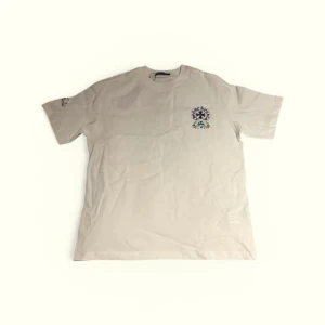 Vit t-shirt från Chrome Hearts - Säljer en vit t-shirt från Chrome Hearts med färgglada blommor och stort tryck på ryggen. T-shirten har korta ärmar och är gjord i bomull. Perfekt för dig som gillar unika och stilrena plagg. Osäker om tröjan är äkta därav priset