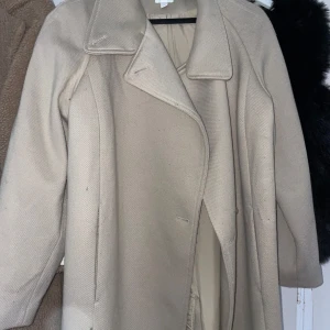 Beige kappa - Elegant beige kappa med klassisk design och breda slag. Perfekt för kyligare dagar med sin långa passform och stilrena look. Kappan har en diskret knäppning framtill och är helfodrad för extra komfort.