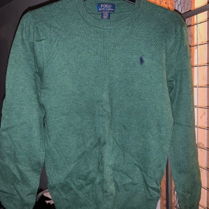 Grön tröja från Polo Ralph Lauren - Säljer en stilren grön tröja från Polo Ralph Lauren i mjuk pima bomull. Tröjan har en klassisk rund halsringning och det ikoniska broderade logotypen på bröstet. Perfekt för en avslappnad och snygg look. Barnstorlek 