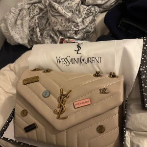 Beige axelväska från Yves Saint Laurent - Snygg beige axelväska från Yves Saint Laurent med quiltad design och guldfärgade kedjedetaljer. Väskan har flera dekorativa märken och ett ikoniskt YSL-emblem på framsidan. Perfekt för att ge din outfit en lyxig touch.