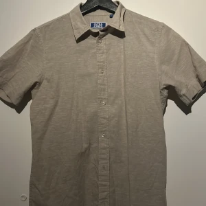 Beige kortärmad skjorta från Jack & Jones - Snygg beige skjorta från Jack & Jones med korta ärmar och klassisk krage. Perfekt för en avslappnad stil. Skjortan har knappar framtill och är tillverkad i ett bekvämt material.