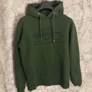 GANT tröja - Säljer en snygg grön hoodie från GANT med justerbar huva och ribbade muddar. Perfekt för en avslappnad stil. Den har ett tryck på framsidan med texten 'GANT American Sportswear'.