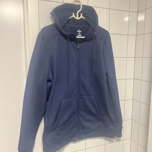 Blå hoodie från Under Armour - Säljer en snygg blå hoodie från Under Armour med dragkedja och justerbar huva. Perfekt för en avslappnad stil. Den har två praktiska fickor framtill och är tillverkad i ett mjukt material.