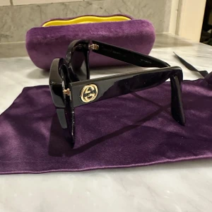 Svarta solglasögon från Gucci - Snygga svarta solglasögon från Gucci med en fyrkantig form. De har en elegant design med märkets logotyp på skalmarna. Kommer med ett lyxigt lila fodral med gul insida för säker förvaring.