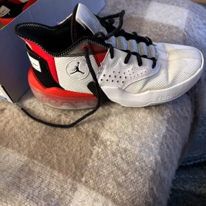 Jordan Basketbollskor Zoom och React - Snygga basketbollskor från Jordan i vitt, svart och rött. Skorna har snörning och är utrustade med Zoom i framfoten och React i hälen för optimal komfort och prestanda. Perfekta för basketentusiaster som vill ha stil och funktion.