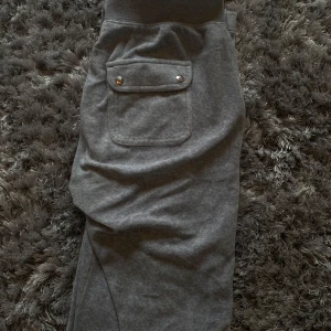 Grå mjukisbyxor från Juicy Couture - Snygga grå mjukisbyxor från Juicy Couture med bekväm resårmidja och snörning. Byxorna har praktiska fickor med knappdetaljer och är perfekta för en avslappnad stil.