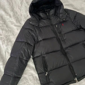 Svart dunjacka från Ralph Lauren - Säljer en snygg svart dunjacka från Ralph Lauren med huva och dragkedja. Jackan har en klassisk design med två fickor framtill och en liten röd logga på bröstet. Perfekt för kyliga dagar! 