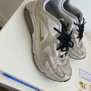 Nike sneakers i beige och guld - Säljer ett par Nike sneakers i beige med guldiga detaljer och svart snörning. Skorna har en unik sula med svarta och guldiga inslag. Perfekta för en sportig look.