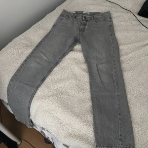 Grå jeans från Jack & Jones - Snygga grå jeans från Jack & Jones med klassisk femficksdesign. De har en dragkedja och knapp framtill. Perfekta för en stilren look.