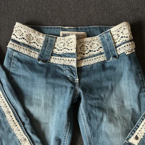 Lågmidjade bootcut med spetsdetaljer - Skitnygga blå bootcut jeans med spetsdetalj vid midjan och längs benen! De är lågmidjade och har en vintage stil. Storleken är 26 och de är uppsprättade nertill för att bli längre! Säljer då de tyvärr inte passar💕🎀