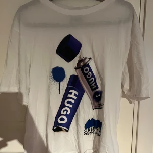 Vit t-shirt med tryck från Hugo - Snygg vit t-shirt från Hugo med ett coolt tryck av sprayburkar i blått och svart. Perfekt för en avslappnad stil med en kreativ touch. T-shirten har korta ärmar och en klassisk passform. Köpt för 1000kr, använd 3-5 gånger.