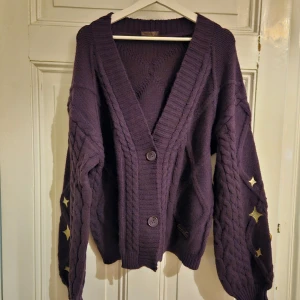 Lila kofta från Speak Now Taylor's Version - OFFICIELL TAYLOR MERCH. Säljer en snygg lila kofta från Speak Now Taylor's Version. Koftan har ett flätstickat mönster och dekorativa stjärnor på ärmarna. Den har en v-ringad design med knappar framtill. Perfekt för att hålla sig varm och stilren! Oversized fit.