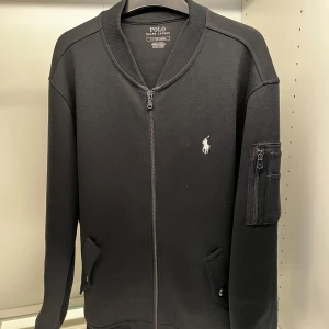 Svart Zip up Polo Ralph Lauren - Snygg svart jacka från Polo Ralph Lauren med dragkedja och en liten broderad logga på bröstet. Jackan har en praktisk ficka med dragkedja på ärmen och två fickor framtill. Perfekt för en stilren look. Endast Swish betalning! Kontakta mig om du har fler frågor!