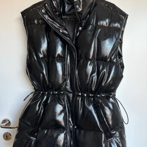 Svart glansig väst H&M - Snygg svart glansig väst med dragkedja och knappar. Västen har en justerbar midja med knytband för en perfekt passform. Perfekt för att hålla värmen med stil. Storlek S