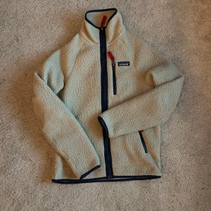 Beige fleecejacka från Patagonia - Mysig beige fleecejacka från Patagonia med dragkedja och hög krage. Jackan har röda detaljer och en praktisk bröstficka med dragkedja. Perfekt för kyliga dagar!
