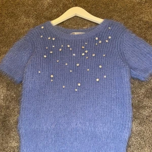 Blå stickad tröja från Zara - Säljer en mysig blå stickad tröja från Zara med korta ärmar och pärldetaljer framtill. Perfekt för att ge en touch av elegans till din outfit. Tröjan har en rund halsringning och är supermjuk.