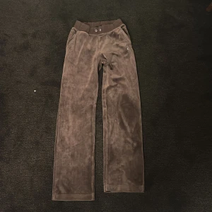 Bruna velourbyxor från Juicy Couture - Snygga bruna velourbyxor från Juicy Couture med bred resår i midjan och logga på baksidan. Byxorna har en avslappnad passform och är perfekta för en bekväm stil.