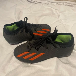 Svarta fotbollsskor från Adidas - Snygga svarta fotbollsskor från Adidas med orangea ränder och grön insida. Skorna har snörning och är perfekta för fotbollsplanen. De har en modern design 