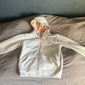 Grå Burberry hoodie med dragkedja - Säljer en grå hoodie med dragkedja och huva. Insidan av huvan har ett rutigt mönster i beige och rött. Tröjan har långa ärmar och är ganska fodrad . Perfekt för en avslappnad stil. Den har några skavanker då på ena armbågen är den sliten som visas på bild samt på båda armändarna så är det slitet med små håll går säker att sy igen😃. Köpte den på Plick 2023 för 1200kr tror den e f@ke burberry! Därav det låga priset. Kan skicka mer bilder 👍