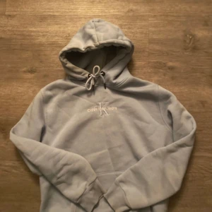 Ljusblå hoodie från Calvin Klein - Säljer en stilren ljusblå hoodie från Calvin Klein Jeans med broderad logga på bröstet. Den har en klassisk design med dragsko i huvan och ribbade muddar vid ärmslut och nederkant. Perfekt för en avslappnad look.