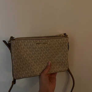 Beige axelväska från Michael Kors - Snygg beige axelväska från Michael Kors med det klassiska MK-mönstret. Väskan har en dragkedja upptill och en justerbar axelrem i brunt läder. Perfekt för att bära dina nödvändigheter med stil.