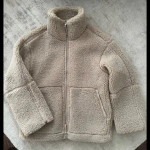 Beige teddyjacka HM - Mysig beige teddyjacka med dragkedja och hög krage. Perfekt för kyliga dagar med sin fluffiga och varma design. Jackan har långa ärmar och två praktiska fickor framtill.