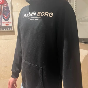 Svart hoodie från Björn Borg - Säljer en svart hoodie från Björn Borgs Autumn/Winter Collection 2021. Tröjan har en klassisk design med en stor ficka framtill och texttryck på bröstet. Perfekt för kyligare dagar.