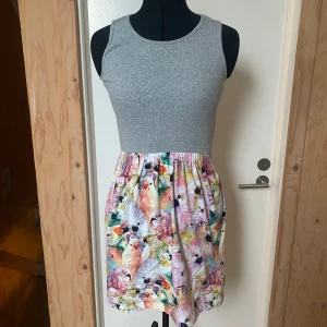 Grå ärmlös klänning med blommigt mönster - Snygg klänning med grå ribbad överdel och färgglatt blommigt mönster på kjolen. Den är ärmlös och perfekt för sommarens varma dagar. Kjolen har en härlig volym och ger en lekfull touch.