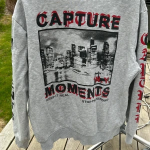 Grå sweatshirt med tryck - Säljer en grå sweatshirt med tryck av en stadssilhuett och texten 'Capture Moments' på baksidan. Texten 'New Yourk' finns på framsidan. Tröjan har långa ärmar och ribbade muddar vid ärmslut och nederkant.