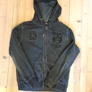 Grå hoodie från Deeluxe - Säljer en grå hoodie från Deeluxe med dragkedja och tryck på bröstet. Tröjan har en justerbar huva med snören och praktiska fickor framtill. Perfekt för en avslappnad stil.