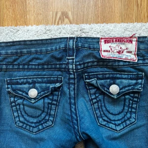 Blå jeans från True Religion - Snygga blå jeans från True Religion med ikoniska bakfickor och dekorativa sömmar. De har en bootcut-stil och är lågmidjade med knappar och dragkedja framtill. Perfekta för en avslappnad look.