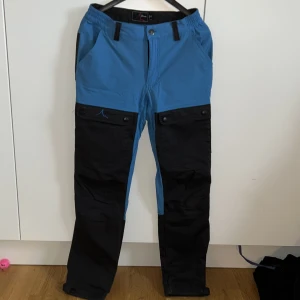 Blå och svarta fritidsbyxor från High Mountain - Snygga och funktionella blå och svarta byxor från High Mountain. De har praktiska fickor med knappar och dragkedja baktill. Perfekta för utomhusaktiviteter med en bekväm passform. De är justerbara längst ner på benen. Byxorna är välanvända och säljer då de inte passar längre.