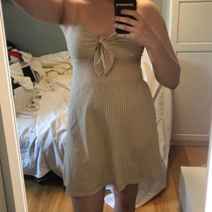 Beige randig klänning Gina tricot - Somrig klänning med knytning vid bysten. Knuten går att ändra på så den kan anpassas för olika storlekar på bysten. Då den är tjock och kan dras åt vid bysten är den lätt att använda utan bh!