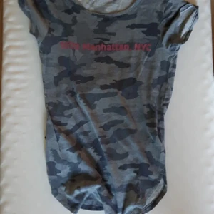 Grå kammo färgad T-shirt i st S - T-shirt i grått kammo mönster med rosa tryck "10173 Manhattan, NYC". St S, jag har vanlogt vis XS men den sitter bra på mig, jag brukar knyta den runt midjan då den är väldigt lång (se bild 2)