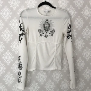 Vit topp med svart tryck - Vit långärmad topp med svart "tatuerings"-tryck från Topshop. Transparent. Väl använd men ändå i bra skick, en aning missfärgad vid armhålorna men inget man tänker på. Går säkert att få bort med något medel. 🤍 Hör av er om ni har några frågor!