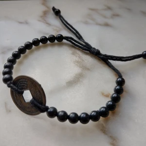 Svart justerbart pärlarmband med metalldetalj - Svart armband med pärlor och en rund metallbricka. Metalldelen är lite rustik och flammig i färgen (mässingsfärgad kan man säga) och med mönster/olika tecken. Pärlorna tror jag är gjorda av plast. Fraktpris 15 kr.