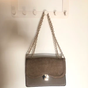 Greige axelremsväska - Grå-Beige-Brun faux mocca-läder väska från H&M med guldigt spänne och kedjeband, använd 1 gång, fint skick. Går att ha både långt och att vika som kort kedjeband. Mått ca:23x7x16 cm (lbh)