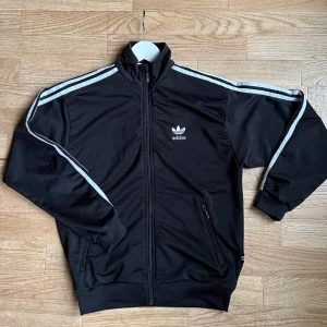 Adidas tracksuit tröja  - 🖤Adidas tröja i använt skick. Super fin och  skön, Helsvart i bak. 100% polyester. passar Xs men inte lika lång i ärmarna. Draget på dragkedjan har en gång lossnat men är lagad, själva dragkedjan funkar oavsett. 
