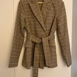 Beigerutig kavaj S - Beigerutig kavaj med skärp som knyts i midjan. Figursydd. Skärpet kan tas bort av om man vill. Använd ett fåtal ggr, i gott skick! Storlek S.