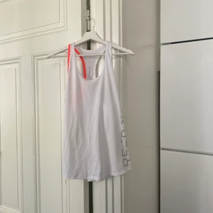 Reebok - träningsslinne - Snyggt, vitt linne med kontrasterande band i orange., Endast använt en gång. Något länge i modellen, halvtransparent. Har legat nerpackat på vinden. Helt och rent. ✨💫