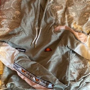 Croppad Ellesse hoodie  - Croppad hoodie från Ellesse. Grå med Ellesse rand på armarna samt lätt rosa och vitt. Skönt material som inte är för varmt! Använd enstaka gång. Strl38 men skulle säga 36!