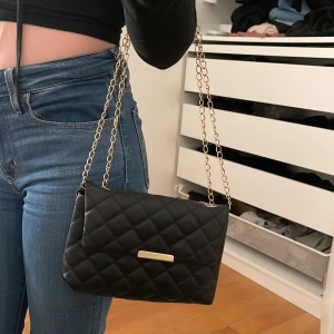 Axelväska  - Svart quiltad axelväska med guldigt spänne framtill 