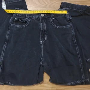 Svarta jeans - Svarta cargoinspirerade jeans med vita sömmar från shein. Hög midja, mått rakt över 31 cm. Broderi på höger bakficka och en byggarbetsbyx-inspirerad tygbit(vet ej vad man ska kalla den??) Skriv för fler bilder eller om frågor uppstår<3