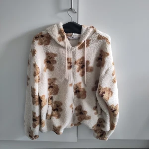Teddyjacka med nallebjörnar  - En beige teddyjacka med bruna nallebjörnar på. Jättesöt och mysig att ha på sig 😊 står ingen tydlig storlek men passar från S-L, den är lite oversized. Köpte den på second hand men är i jättebra skick. Det finns en liten fläck under ena ärmen men märks ej