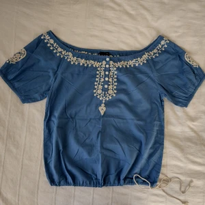 Blå blus/topp/singoallatopp med brodyr  - Blå blus med knappar och brodyr i benvitt/beiget. Dragsko i nederkant. 100% bomull. Använd en gång och i fint skick. Storlek 36. Bred över axlarna och jag skulle tro att blusen kan passa även de som har storlek 38/40. Extraknappar medföljer.