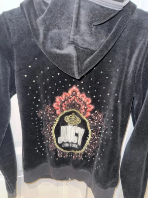 Juicy Couture svart velour hoodie - Snygg svart hoodie från Juicy Couture i mjuk velour med stor broderad logga och krona på ryggen. Hoodien har huva och dekoreras med glittriga stenar och detaljer i rött och guld. Perfekt för dig som gillar statement-plagg och vill sticka ut. Mesta dels av stenar saknas men ser fortfarande jätte fin ut.
