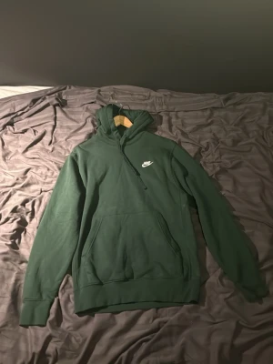 Mörkgrön Nike hoodie med logga - Snygg mörkgrön hoodie från Nike med vit broderad logga på bröstet. Klassisk känguruficka framtill, ribbade muddar och justerbar huva med dragsko. Perfekt för en avslappnad och sportig stil.